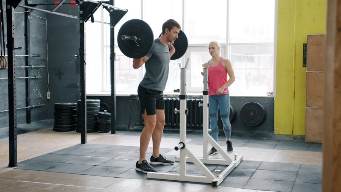 Muscular young man training in gym squatting with barbell working out with coach attractive girl wearing sports clothing. People, workout and leisure concept.