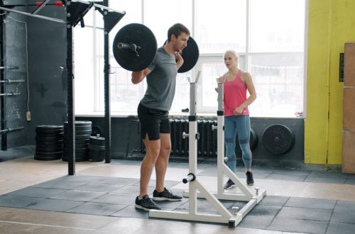 Muscular young man training in gym squatting with barbell working out with coach attractive girl wearing sports clothing. People, workout and leisure concept.